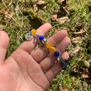 Puede incluir: Una pulsera de cuentas amarillas, azules y plateadas. La pulsera tiene un dije de bot&oacute;n y un dije de lib&eacute;lula. Las cuentas son de varias formas, redondas y cil&iacute;ndricas. La pulsera se sostiene en una mano.