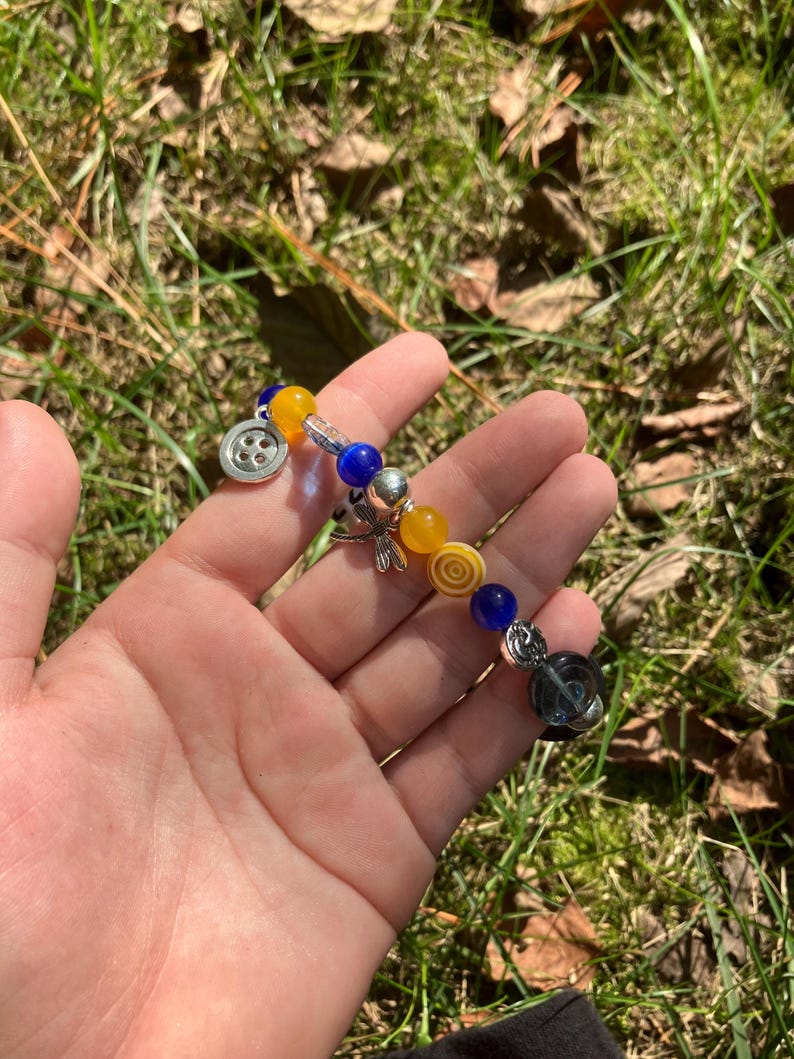 Puede incluir: Una pulsera de cuentas amarillas, azules y plateadas. La pulsera tiene un dije de bot&oacute;n y un dije de lib&eacute;lula. Las cuentas son de varias formas, redondas y cil&iacute;ndricas. La pulsera se sostiene en una mano.