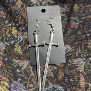 Puede incluir: Un par de pendientes plateados con forma de espada. Cada pendiente presenta un diseño detallado de espada con una hoja larga y delgada y una empuñadura en forma de cruz. Los pendientes se muestran en una tarjeta negra.