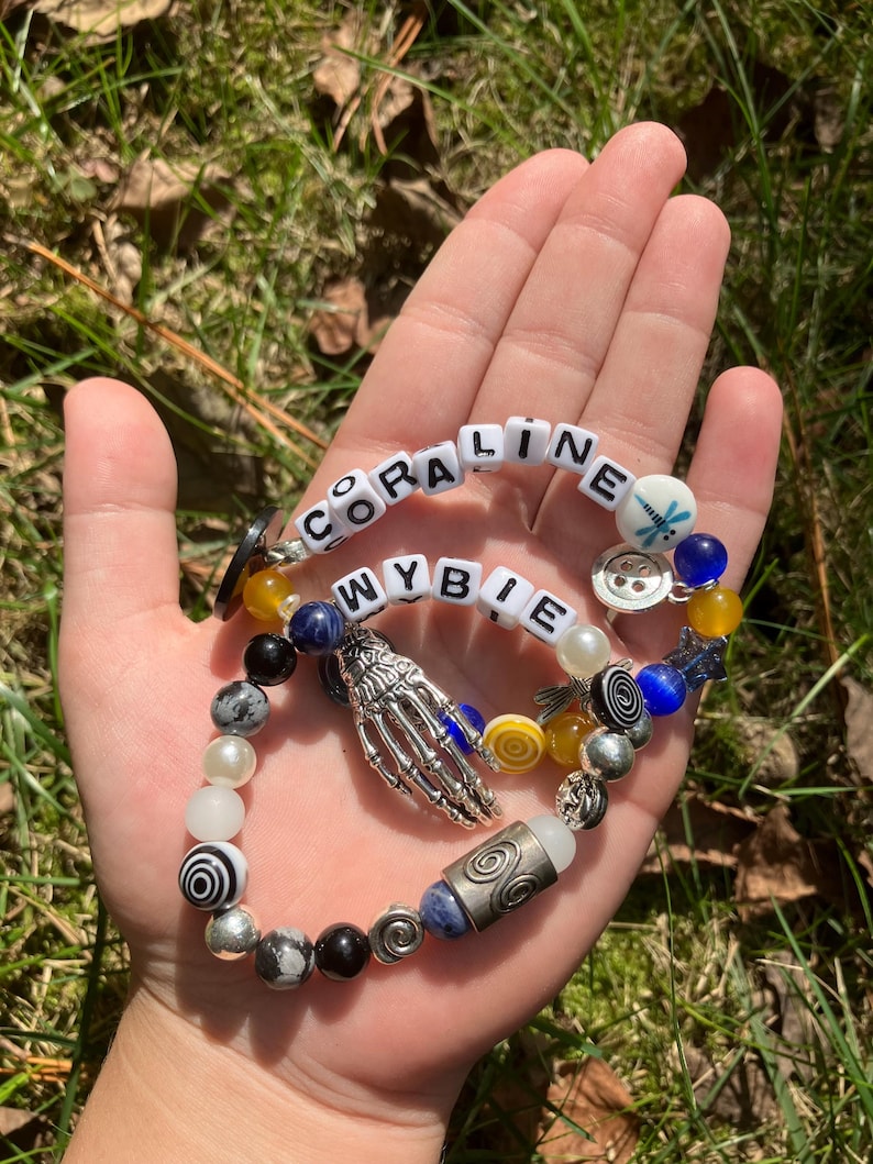 May include: Two beaded bracelets with the names "CORALINE" and "WYBIE" spelled out in white letter beads. The bracelets feature a variety of beads in colors like black, white, blue, and yellow, with a silver skeleton hand charm.
