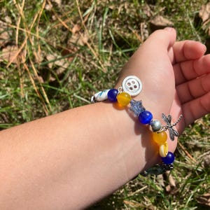 Puede incluir: Una pulsera de cuentas con cuentas azules, amarillas y blancas, y abalorios plateados. La pulsera presenta un dije de lib&eacute;lula y un dije de bot&oacute;n. La pulsera est&aacute; en una mu&ntilde;eca.