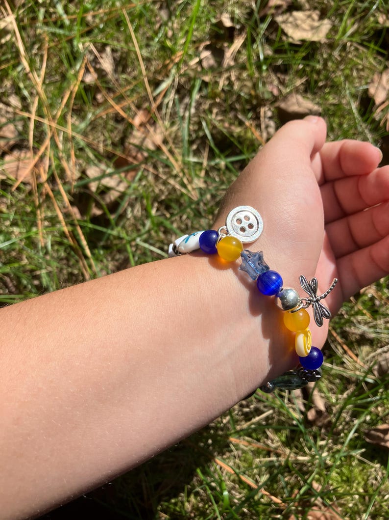 Puede incluir: Una pulsera de cuentas con cuentas azules, amarillas y blancas, y abalorios plateados. La pulsera presenta un dije de lib&eacute;lula y un dije de bot&oacute;n. La pulsera est&aacute; en una mu&ntilde;eca.