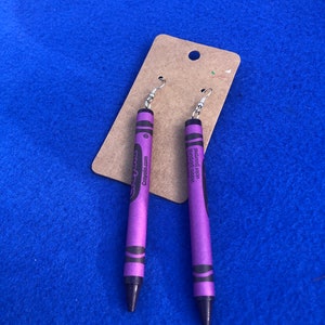 May include: A pair of purple crayon earrings with silver hooks. The crayons have the Crayola logo and the words "Violet Purple" printed on them.