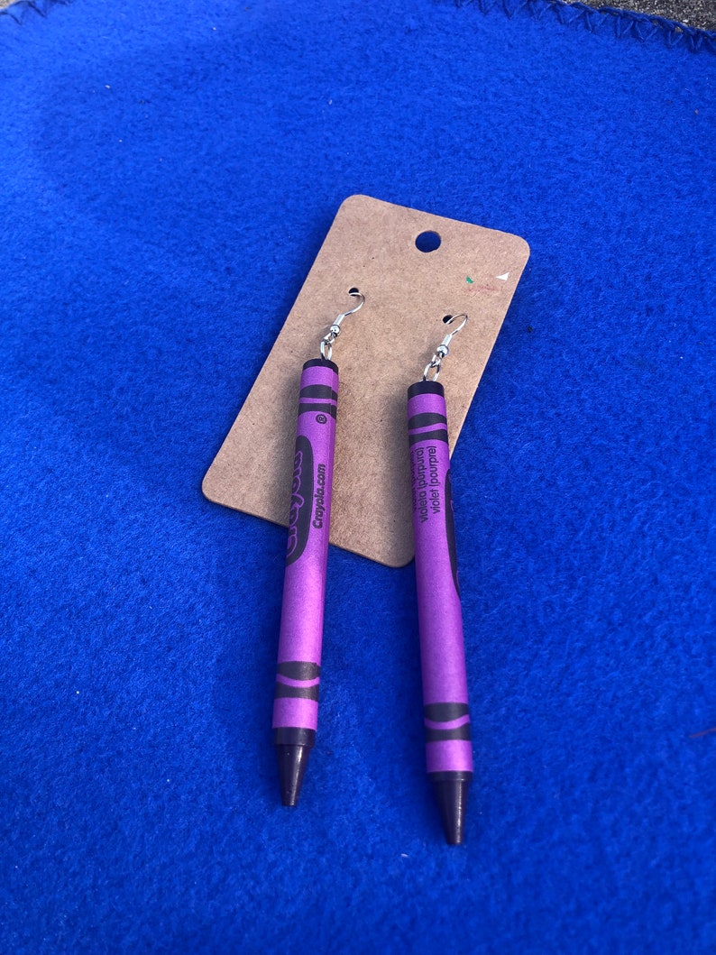 May include: A pair of purple crayon earrings with silver hooks. The crayons have the Crayola logo and the words "Violet Purple" printed on them.