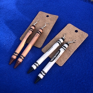 May include: A pair of brown crayon earrings and a pair of white crayon earrings with black tips. The earrings are on a brown card with the text "Crayola.com" printed on it.