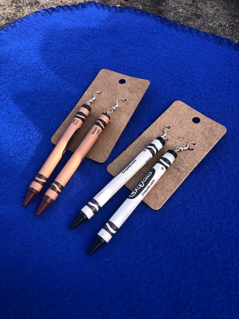 May include: A pair of brown crayon earrings and a pair of white crayon earrings with black tips. The earrings are on a brown card with the text "Crayola.com" printed on it.