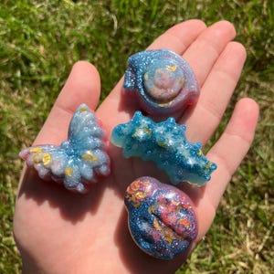 Puede incluir: Cuatro pequeñas esculturas de resina coloridas con detalles de purpurina. Las esculturas son en tonos azules, rosas y dorados, y tienen forma de criaturas marinas. Los objetos se sostienen en una mano abierta sobre un fondo de hierba verde.