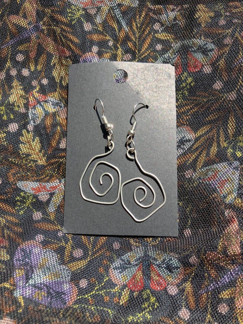 K&ouml;nnte beinhalten: Silberfarbene Drahtohrringe mit Spiraldesign, pr&auml;sentiert auf einer schwarzen Karte. Die Ohrringe haben einen Hakenverschluss. Der Hintergrund ist ein dunkler Stoff mit floralem Muster.