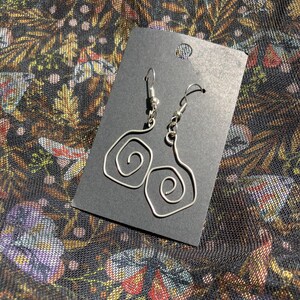 K&ouml;nnte beinhalten: Silberfarbene Drahtohrringe mit Spiraldesign, pr&auml;sentiert auf einer schwarzen Karte. Die Ohrringe haben einen Hakenverschluss. Der Hintergrund ist ein dunkler Stoff mit einem Blumenmuster in Gold-, Rot- und Lilat&ouml;nen.