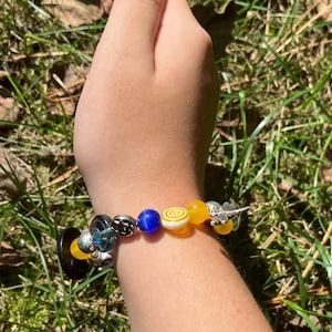 Puede incluir: Una pulsera de cuentas con una variedad de cuentas coloridas, incluyendo amarillas, azules y de vidrio transparente. La pulsera presenta un dije de lib&eacute;lula plateada y una cuenta negra en forma de bot&oacute;n. La pulsera se lleva en la mu&ntilde;eca.