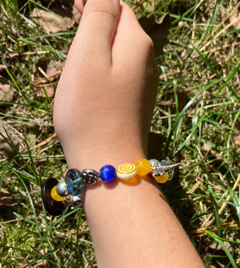 Puede incluir: Una pulsera de cuentas con una variedad de cuentas coloridas, incluyendo amarillas, azules y de vidrio transparente. La pulsera presenta un dije de lib&eacute;lula plateada y una cuenta negra en forma de bot&oacute;n. La pulsera se lleva en la mu&ntilde;eca.