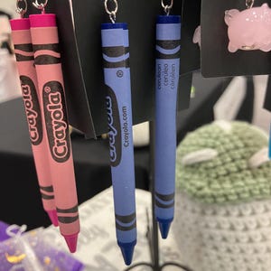 May include: A pair of pink and blue Crayola crayon earrings. The pink crayon is labeled "Crayola" and the blue crayon is labeled "Crayola" and "Crayola.com". The blue crayon also has the word "cerulean" written on it in French, Spanish, and English.