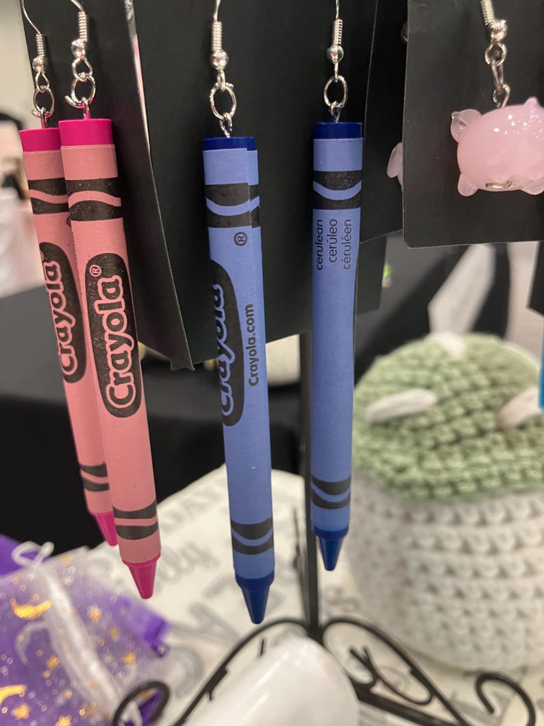 May include: A pair of pink and blue Crayola crayon earrings. The pink crayon is labeled "Crayola" and the blue crayon is labeled "Crayola" and "Crayola.com". The blue crayon also has the word "cerulean" written on it in French, Spanish, and English.