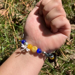Puede incluir: Una pulsera de cuentas con cuentas amarillas, azules y plateadas. La pulsera incluye un dije de lib&eacute;lula plateada y un cierre negro y amarillo. La pulsera se muestra en una mu&ntilde;eca sobre un fondo de hierba verde y hojas.