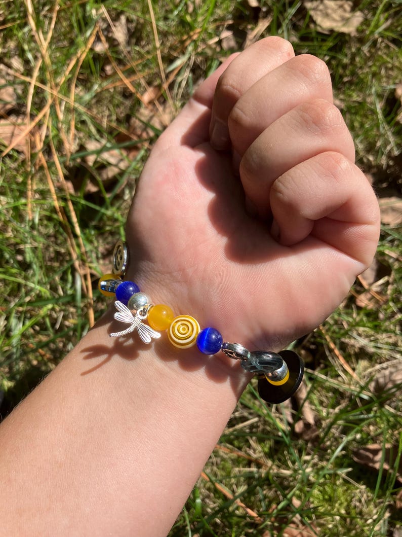 Puede incluir: Una pulsera de cuentas con cuentas amarillas, azules y plateadas. La pulsera incluye un dije de lib&eacute;lula plateada y un cierre negro y amarillo. La pulsera se muestra en una mu&ntilde;eca sobre un fondo de hierba verde y hojas.