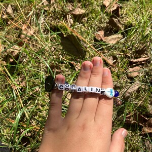 Puede incluir: Una pulsera de cuentas blancas que deletrean "CORALINE". La pulsera tiene un cierre de bot&oacute;n negro y un dije azul y blanco. La pulsera se sostiene sobre un fondo de hierba verde.