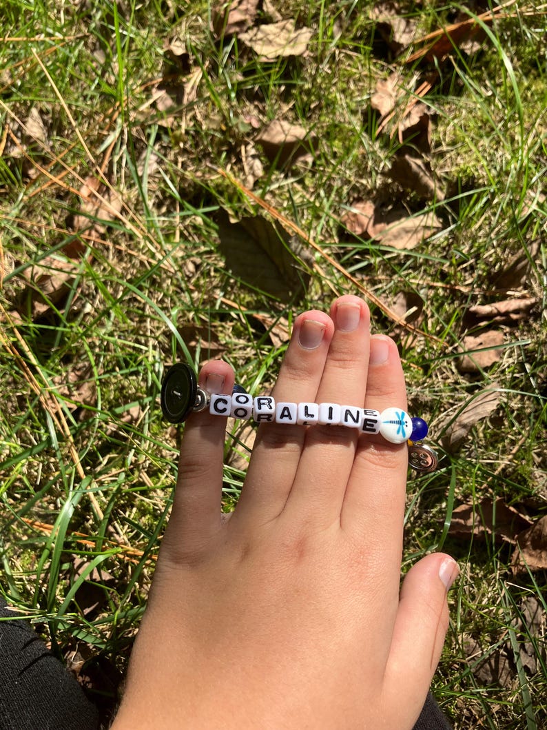 Puede incluir: Una pulsera de cuentas blancas que deletrean "CORALINE". La pulsera tiene un cierre de bot&oacute;n negro y un dije azul y blanco. La pulsera se sostiene sobre un fondo de hierba verde.