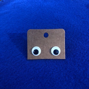 May include: A pair of googly eyes on a brown card. The eyes are white with black pupils.
