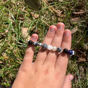 May include: A beaded bracelet featuring a variety of stone beads in black, white, gray, and blue. The bracelet is held in a hand against a backdrop of green grass and sunlight.