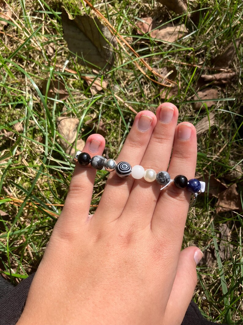 May include: A beaded bracelet featuring a variety of stone beads in black, white, gray, and blue. The bracelet is held in a hand against a backdrop of green grass and sunlight.