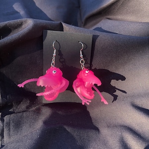 May include: A pair of pink monster earrings with silver hooks. The monsters have open mouths and are made of a translucent material.