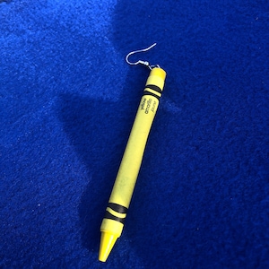 May include: A yellow crayon earring with a silver hook. The crayon is labeled "yellow" and "amarillo".