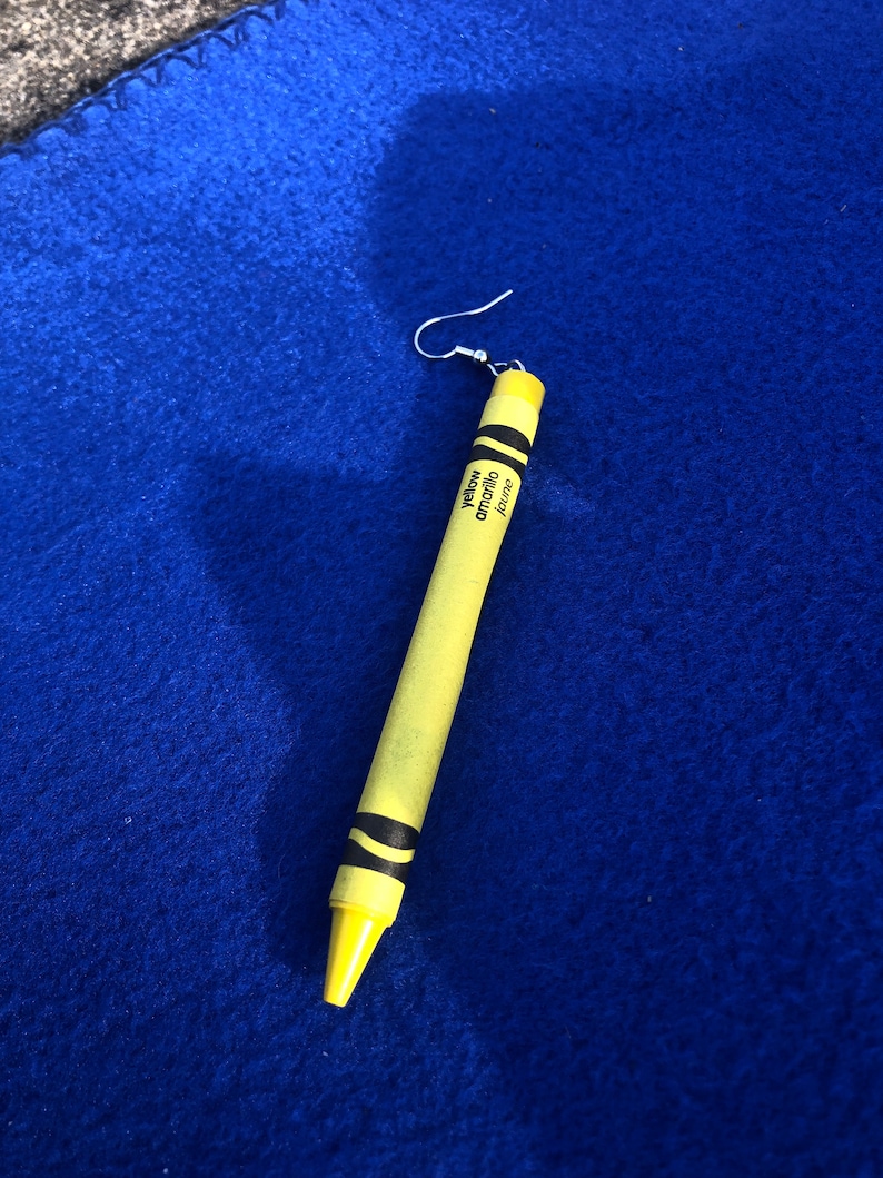 May include: A yellow crayon earring with a silver hook. The crayon is labeled "yellow" and "amarillo".