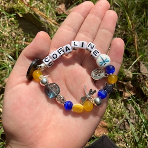 Puede incluir: Una pulsera hecha a mano con el nombre "CORALINE" escrito con cuentas de letras blancas. La pulsera presenta cuentas azules, amarillas y plateadas, adem&aacute;s de dijes como una lib&eacute;lula y un bot&oacute;n. La pulsera se muestra en una mano abierta.
