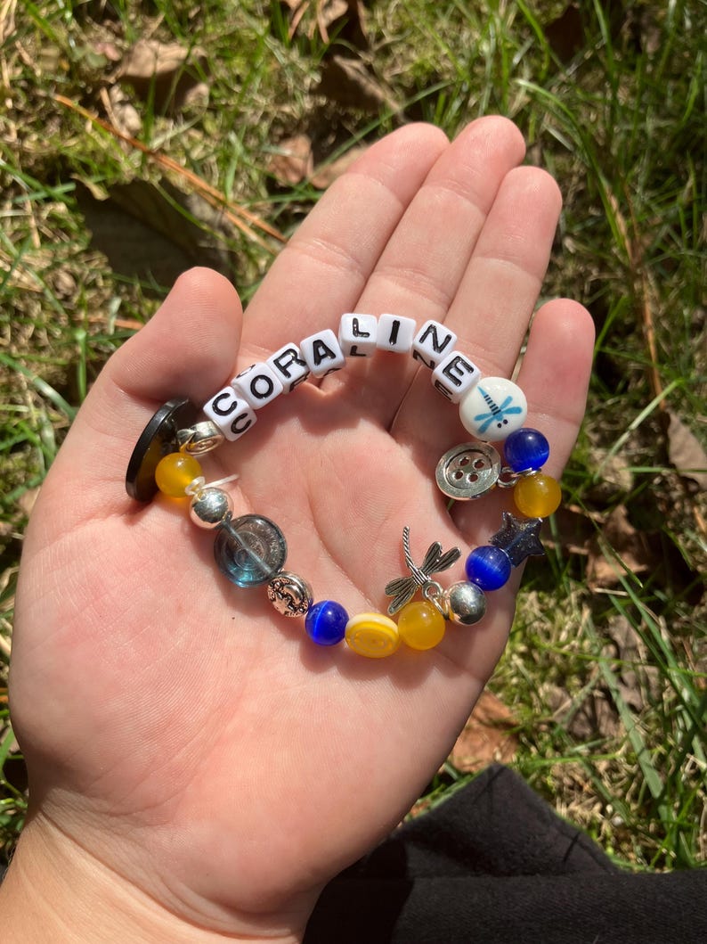 Puede incluir: Una pulsera hecha a mano con el nombre "CORALINE" escrito con cuentas de letras blancas. La pulsera presenta cuentas azules, amarillas y plateadas, adem&aacute;s de dijes como una lib&eacute;lula y un bot&oacute;n. La pulsera se muestra en una mano abierta.