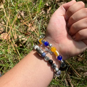 May include: Two beaded bracelets with a variety of beads in silver, black, blue, yellow, and white. One bracelet features a dragonfly charm. The bracelets are worn on a wrist against a background of green grass.