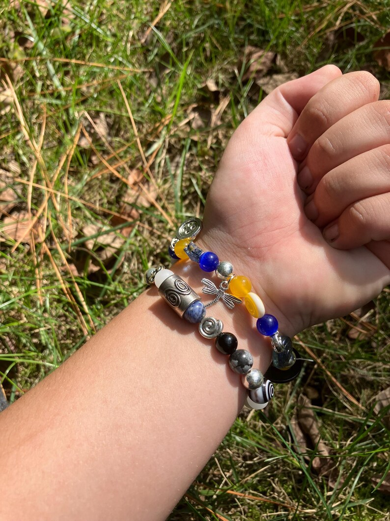 May include: Two beaded bracelets with a variety of beads in silver, black, blue, yellow, and white. One bracelet features a dragonfly charm. The bracelets are worn on a wrist against a background of green grass.