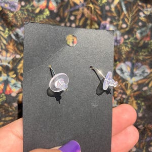K&ouml;nnte beinhalten: Ein Paar silberfarbene Ohrstecker mit transparenten Kunststoffverschl&uuml;ssen, pr&auml;sentiert auf einer schwarzen Karte. Die Ohrringe werden von hinten betrachtet, wobei die Stifte und Verschl&uuml;sse sichtbar sind. Der Hintergrund zeigt ein farbenfrohes Blumenmuster.