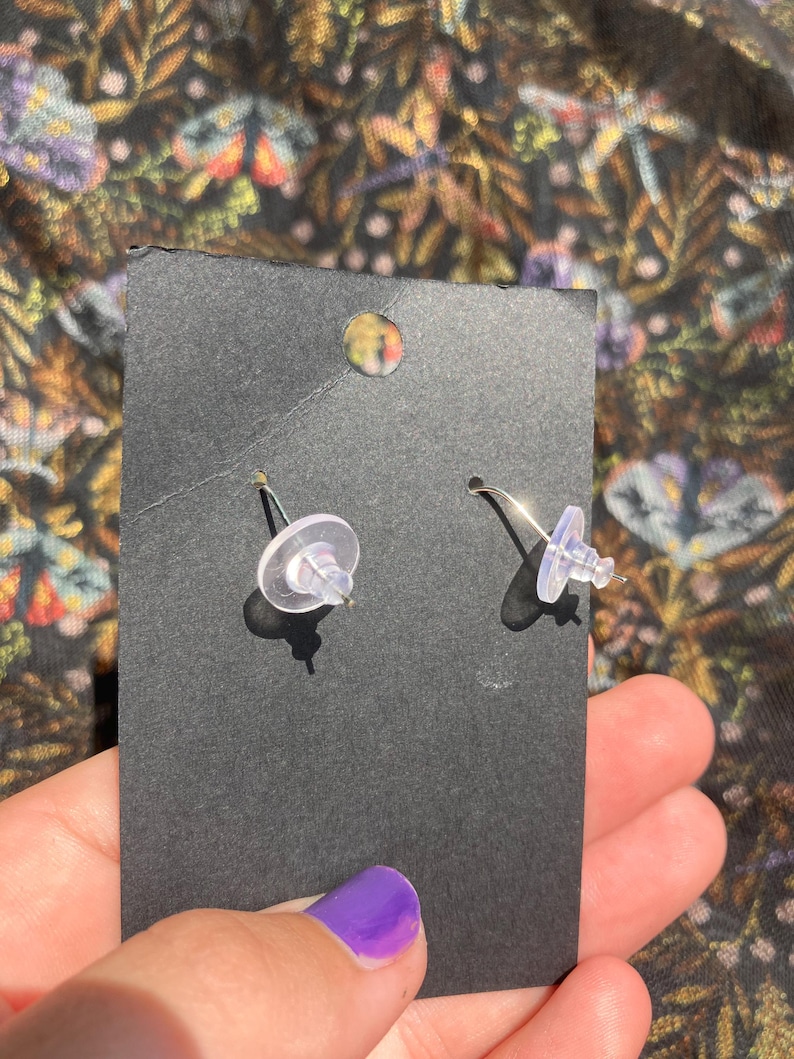 K&ouml;nnte beinhalten: Ein Paar silberfarbene Ohrstecker mit transparenten Kunststoffverschl&uuml;ssen, pr&auml;sentiert auf einer schwarzen Karte. Die Ohrringe werden von hinten betrachtet, wobei die Stifte und Verschl&uuml;sse sichtbar sind. Der Hintergrund zeigt ein farbenfrohes Blumenmuster.
