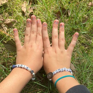 May include: Two beaded bracelets spelling out "WYBIE" and "CORALINE" are displayed. The "WYBIE" bracelet features blue beads and a pearl, while the "CORALINE" bracelet is on a turquoise cord. Both bracelets are on a wrist.