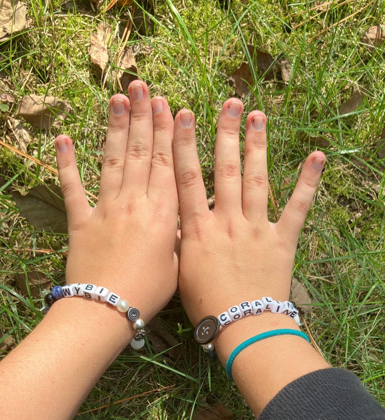 May include: Two beaded bracelets spelling out "WYBIE" and "CORALINE" are displayed. The "WYBIE" bracelet features blue beads and a pearl, while the "CORALINE" bracelet is on a turquoise cord. Both bracelets are on a wrist.