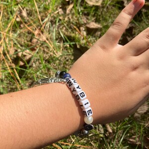 May include: A beaded bracelet spelling "WYBIE" with white letter beads. The bracelet includes a mix of blue, black, and white beads, and a silver-toned charm. The bracelet is worn on a wrist.