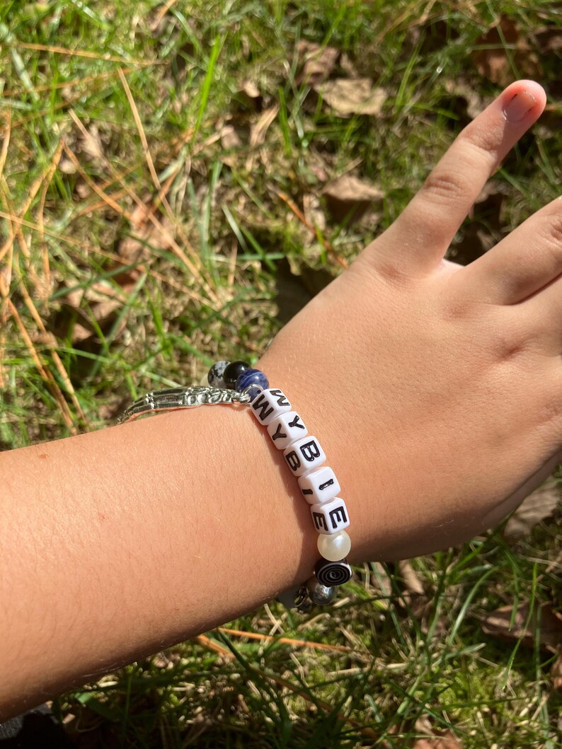 May include: A beaded bracelet spelling "WYBIE" with white letter beads. The bracelet includes a mix of blue, black, and white beads, and a silver-toned charm. The bracelet is worn on a wrist.