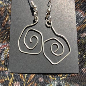 K&ouml;nnte beinhalten: Ein Paar silberfarbene Drahtohrringe. Jeder Ohrring hat ein Spiraldesign und einen Hakenverschluss. Die Ohrringe werden auf einer schwarzen Oberfl&auml;che vor einem gemusterten Hintergrund gezeigt.