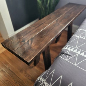 May include: A dark brown wooden bedside table with a simple design. The table has a rectangular top and two angled legs on each side. The table is made of unfinished wood and has a natural wood grain.