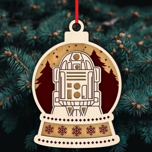 K&ouml;nnte beinhalten: Weihnachtsbaumschmuck aus Holz mit R2-D2 aus Star Wars. Der Schmuck hat die Form einer Schneekugel mit rotem Hintergrund und einer Waldsilhouette. Die Basis hat ein Schneeflockenmuster und ein rotes Band zum Aufh&auml;ngen.