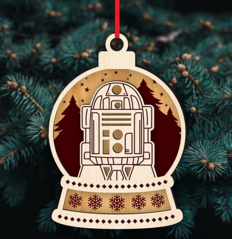K&ouml;nnte beinhalten: Weihnachtsbaumschmuck aus Holz mit R2-D2 aus Star Wars. Der Schmuck hat die Form einer Schneekugel mit rotem Hintergrund und einer Waldsilhouette. Die Basis hat ein Schneeflockenmuster und ein rotes Band zum Aufh&auml;ngen.