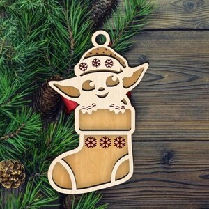 K&ouml;nnte beinhalten: Weihnachtsschmuck aus Holz mit einer Figur in einem Strumpf und Hut. Der Schmuck ist hellbraun mit Schneeflocken-Ausschnitten und einer roten Schleife. Der Hintergrund umfasst Tannennadeln und eine Holzoberfl&auml;che.
