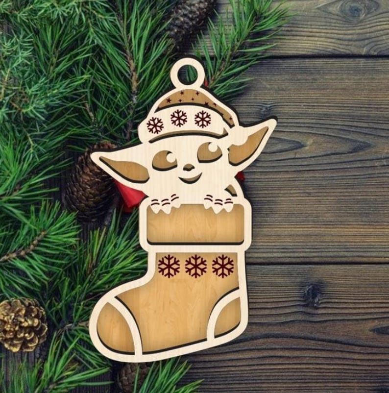 K&ouml;nnte beinhalten: Weihnachtsschmuck aus Holz mit einer Figur in einem Strumpf und Hut. Der Schmuck ist hellbraun mit Schneeflocken-Ausschnitten und einer roten Schleife. Der Hintergrund umfasst Tannennadeln und eine Holzoberfl&auml;che.