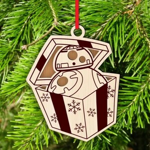 K&ouml;nnte beinhalten: Weihnachtsbaumschmuck aus Holz mit einem BB-8-Droiden in einer Geschenkbox. Die Box ist mit Schneeflocken und dunkelroten Streifen verziert. Der Schmuck h&auml;ngt an einem roten Band.