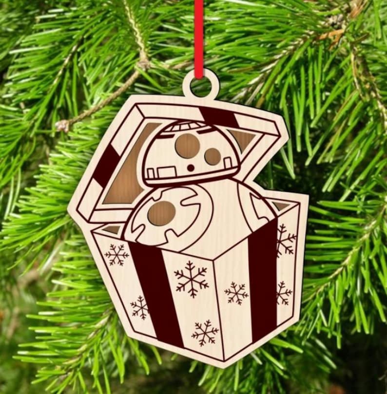 K&ouml;nnte beinhalten: Weihnachtsbaumschmuck aus Holz mit einem BB-8-Droiden in einer Geschenkbox. Die Box ist mit Schneeflocken und dunkelroten Streifen verziert. Der Schmuck h&auml;ngt an einem roten Band.