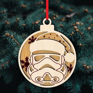 K&ouml;nnte beinhalten: Weihnachtsbaumschmuck aus Holz mit einem Stormtrooper-Helm, der einen Weihnachtsmannhut tr&auml;gt. Der runde Schmuck hat ein rotes Band zum Aufh&auml;ngen. Schneeflocken- und Sterndetails sind in das Holz geschnitten.
