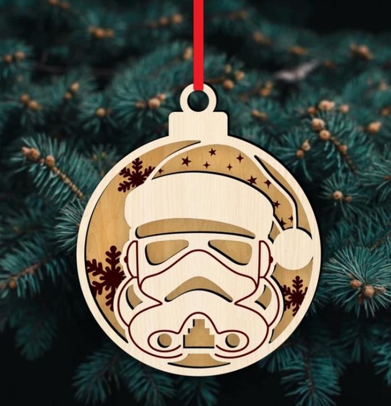 K&ouml;nnte beinhalten: Weihnachtsbaumschmuck aus Holz mit einem Stormtrooper-Helm, der einen Weihnachtsmannhut tr&auml;gt. Der runde Schmuck hat ein rotes Band zum Aufh&auml;ngen. Schneeflocken- und Sterndetails sind in das Holz geschnitten.