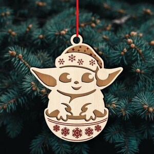 K&ouml;nnte beinhalten: Weihnachtsbaumschmuck aus Holz mit einer stilisierten Figur mit Weihnachtsm&uuml;tze und Schneeflockenmuster. Der Anh&auml;nger h&auml;ngt an einem roten Faden vor einem Hintergrund aus Tannenzweigen. Die Figur befindet sich in einer Sch&uuml;ssel mit Schneeflockendetails.