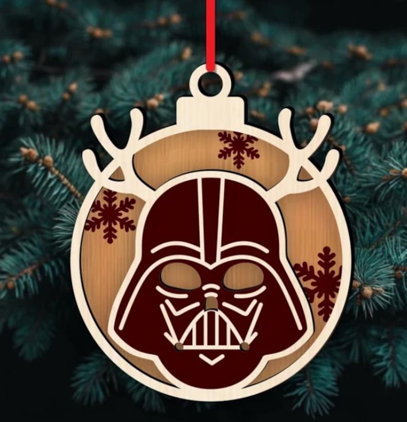 K&ouml;nnte beinhalten: Weihnachtsschmuck aus Holz mit Darth Vaders Helmdesign. Der Schmuck hat eine hellbraune Holzbasis mit einer dunkelroten Helmumrandung und Schneeflocken-Akzenten. Ein rotes Band ist zum Aufh&auml;ngen befestigt.