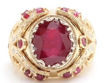 May include: A gold ring featuring a large, oval-cut ruby as the centerpiece. The ring is adorned with smaller rubies and diamonds, set within an ornate, filigree design. The ring's band and setting are a warm gold color.
